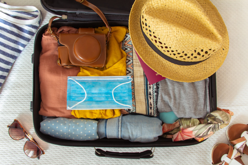 Valise ouverte soigneusement remplie de vêtements pliés, avec un chapeau de paille, un appareil photo, des lunettes de soleil et un masque disposés sur le dessus.