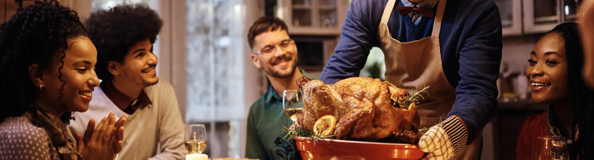 group of five friends at friendsgiving dinner while man sets turkey on table