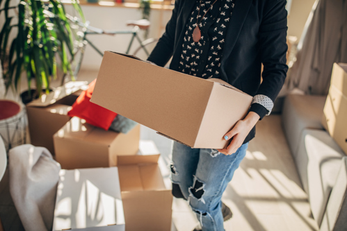 person carrying a cardboard box in a living room surrounded by moving boxes