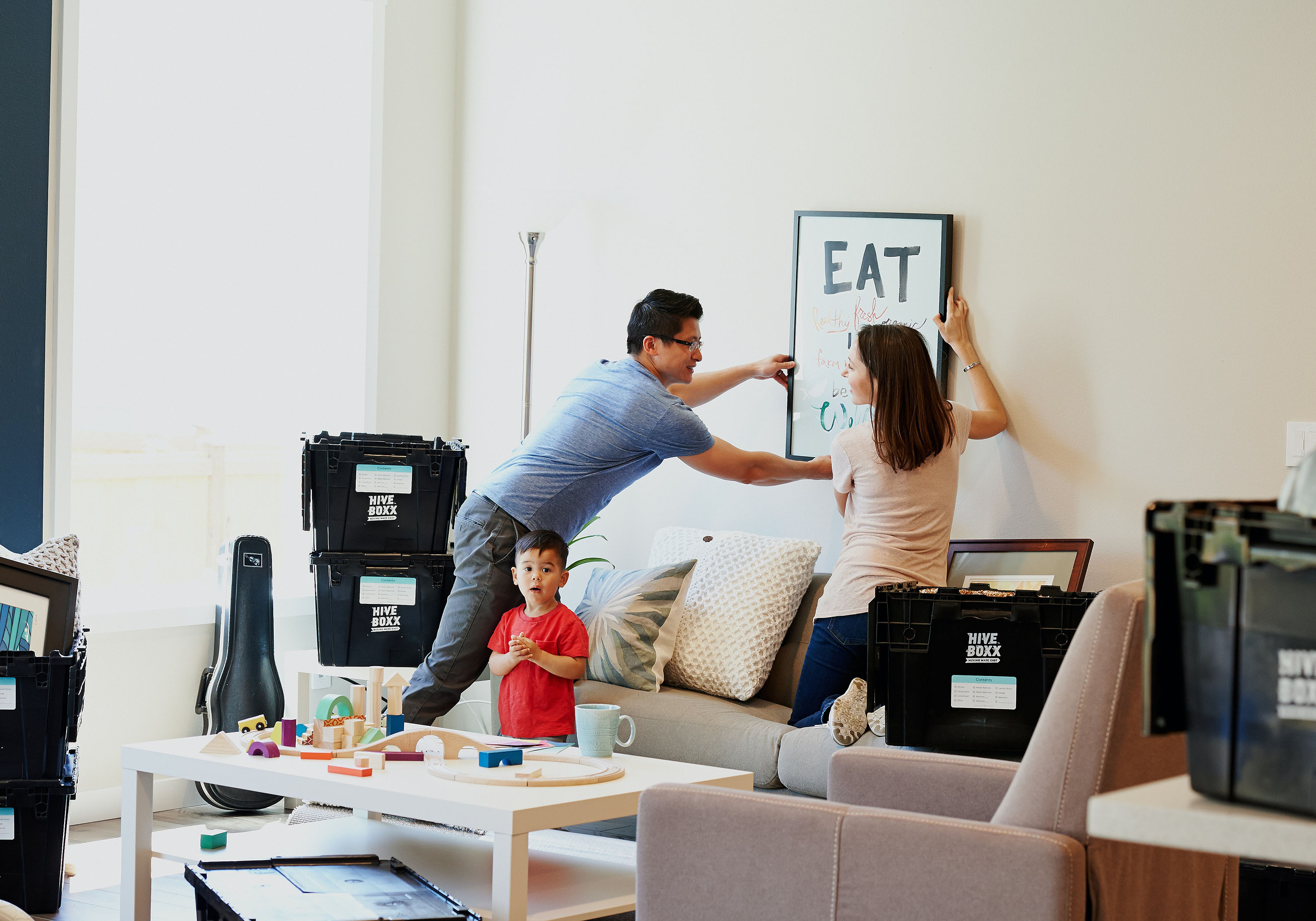 parents hanging a framed art piece in a new home, surrounded by moving boxes, with a child behind them looking at the camera.