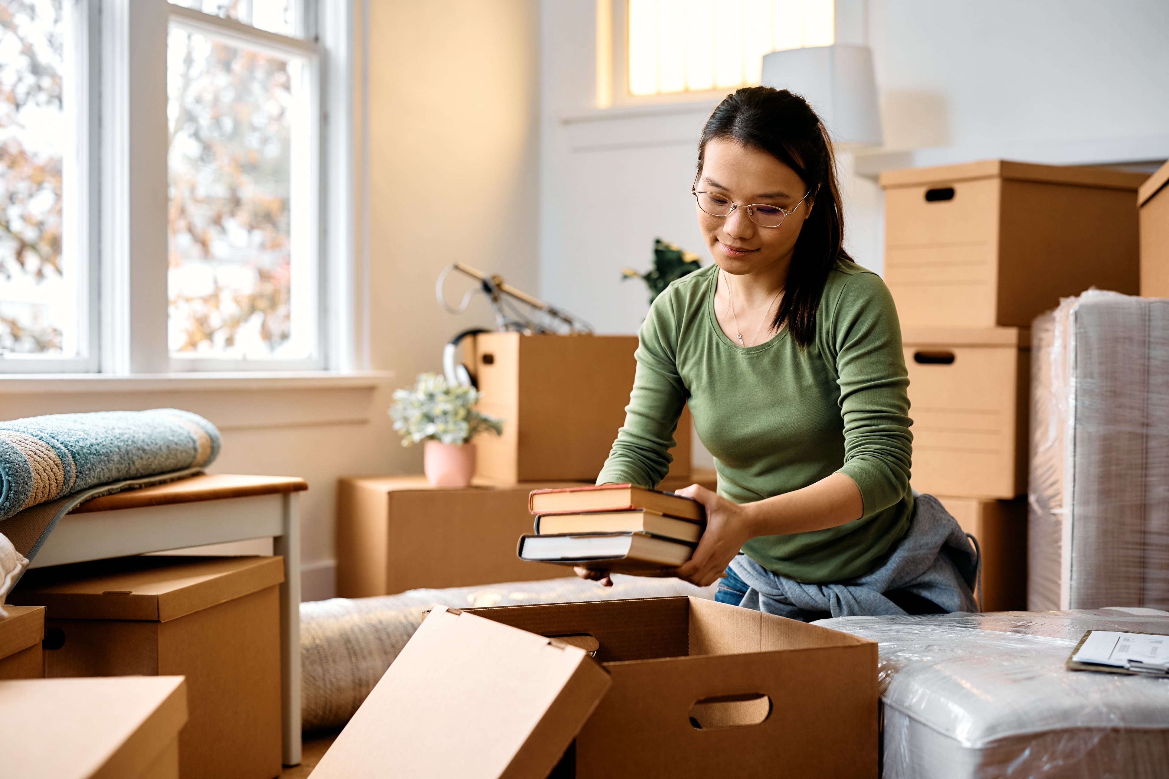 woman with glasses surrounded by moving boxes packing up a handful of books