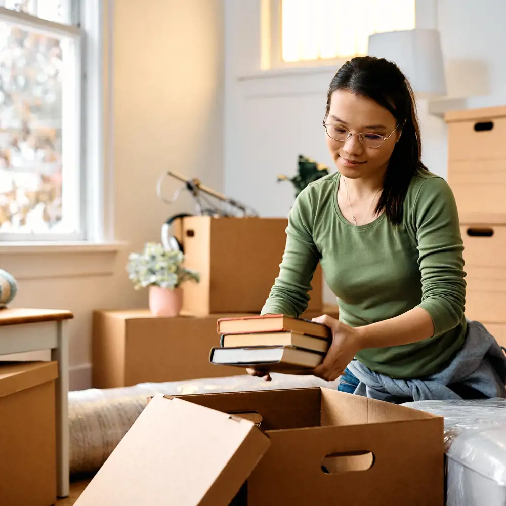 woman with glasses surrounded by moving boxes packing up a handful of books