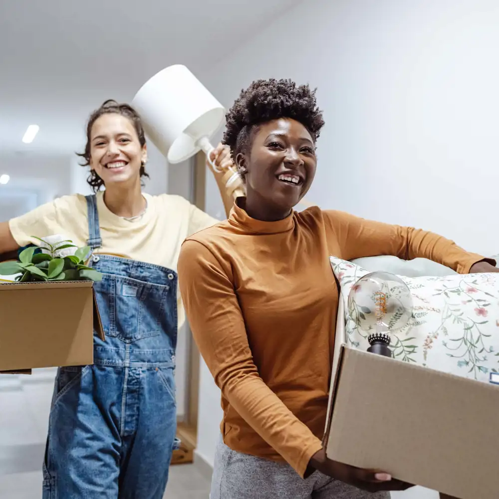two college students smiling as they walk through dorm hallway with boxes of items