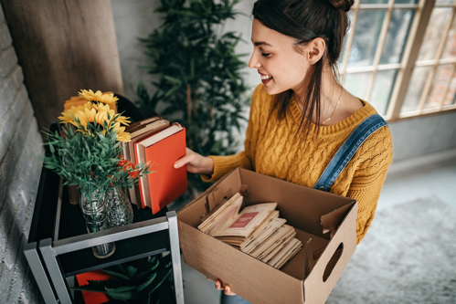 femme plaçant des livres sur une étagère tout en tenant une boîte d’objets dans un espace de vie chaleureux