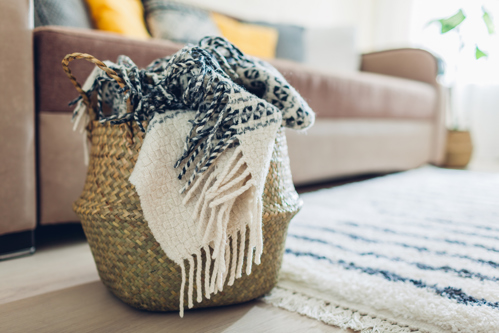 panier tressé rempli de couvertures et d’une écharpe à côté d’un canapé et d’un tapis