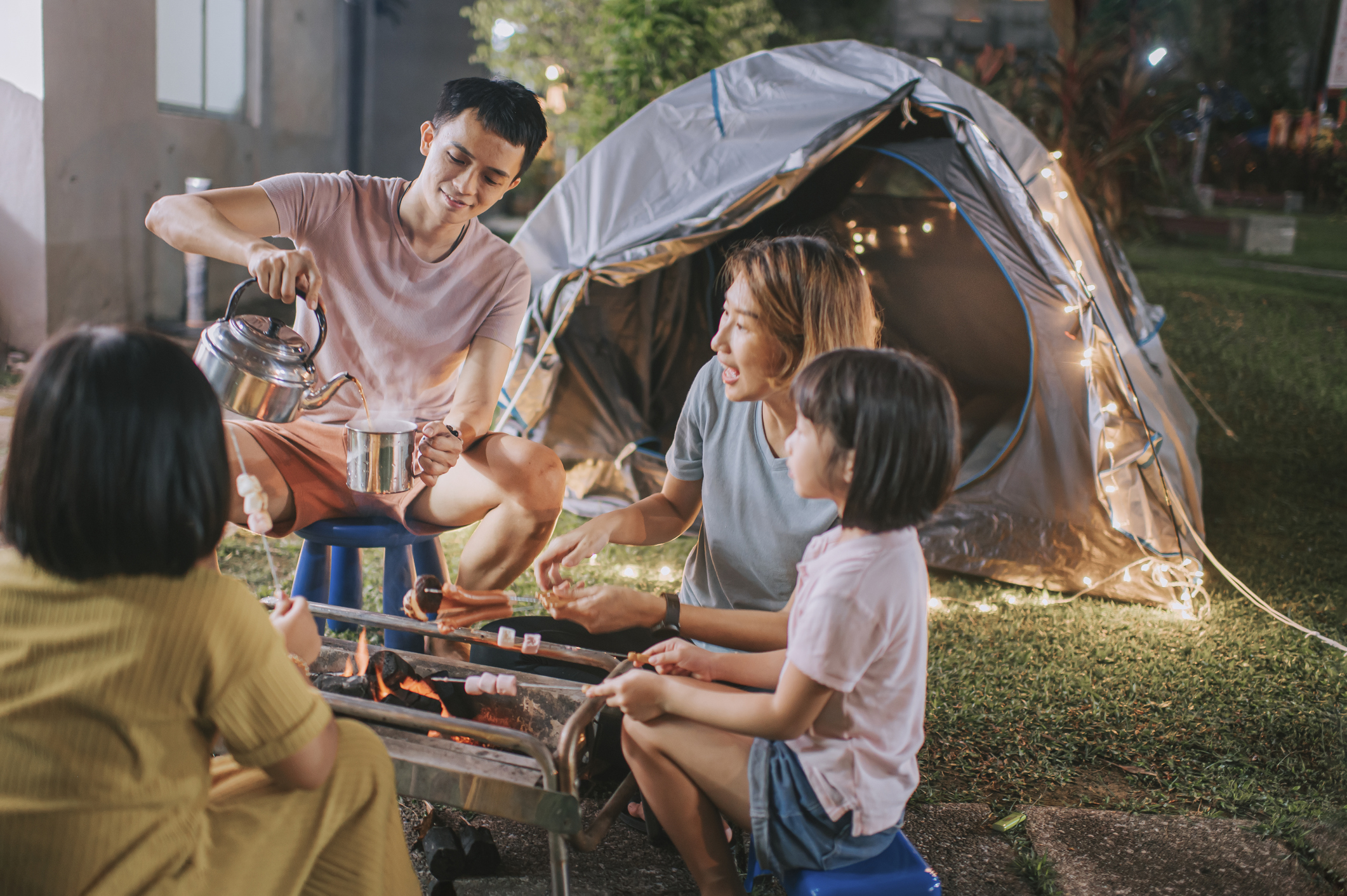 Une famille avec deux jeunes enfants fait du camping dans son jardin avec des grillades et des guirlandes électriques.