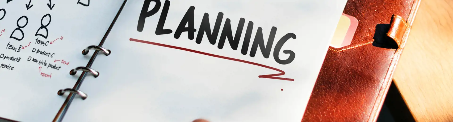 Hand gripping pen above notebook with the word planning written in black ink and underlined with red ink on white page.