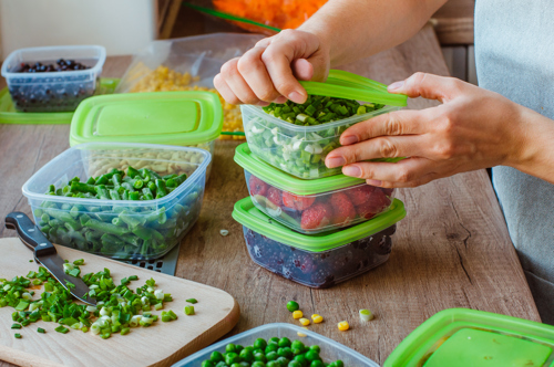 person stacking green-lidded containers filled with chopped vegetables on a kitchen counter
