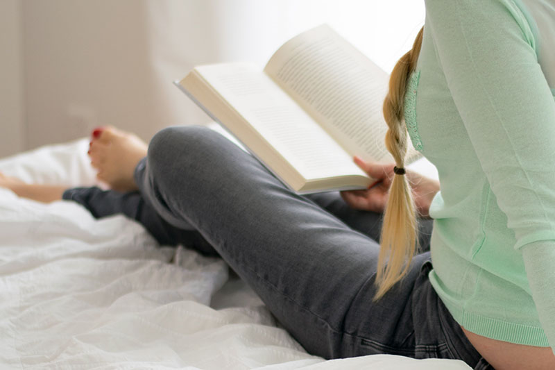 A student sits on her bed reading a book.