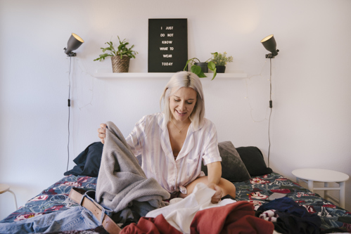 woman sitting on bed sorting through a pile of clothes in a bright bedroom
