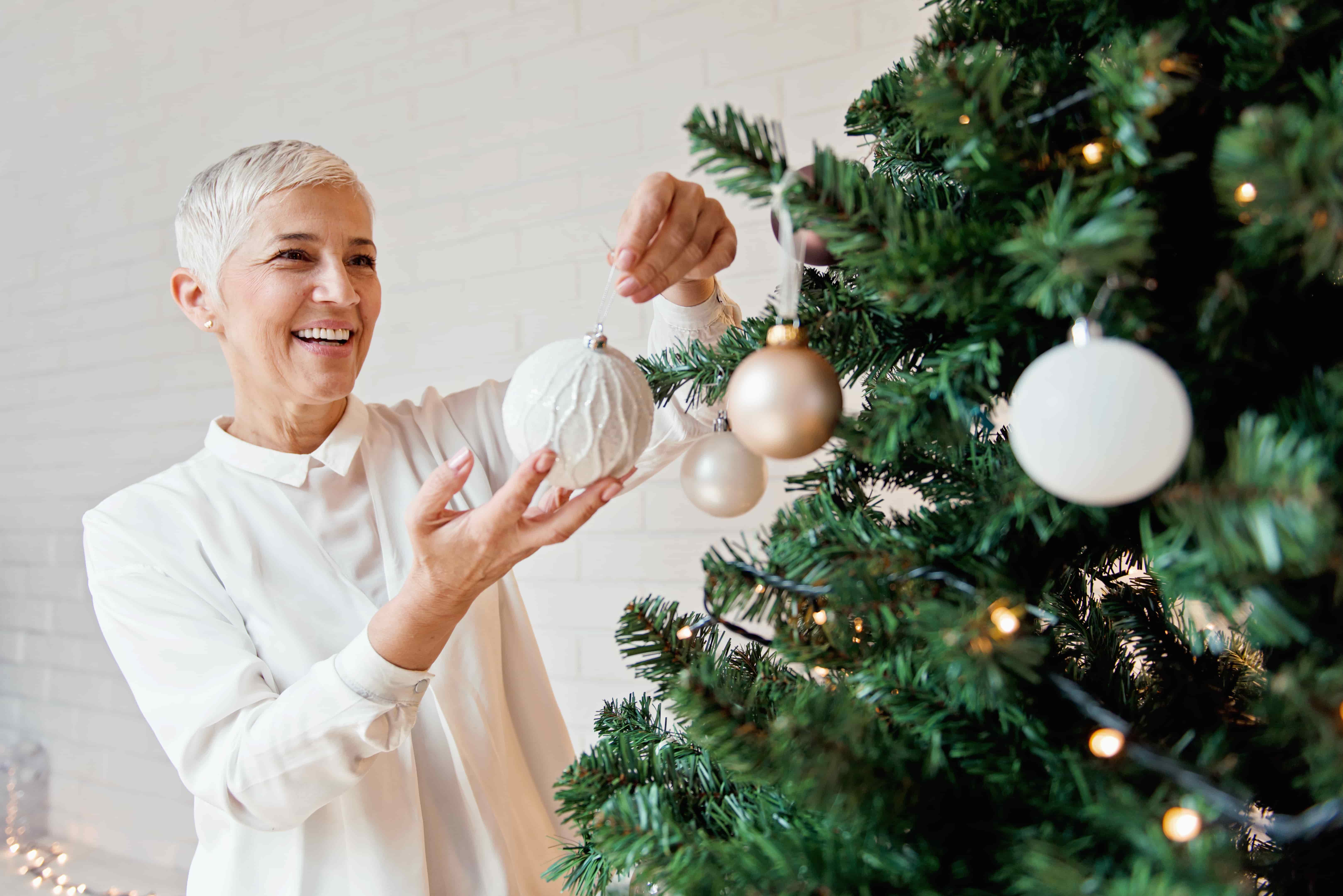 older woman adding white bauble ornament to christmas tree