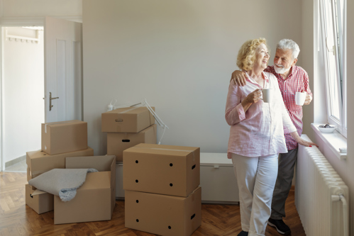 elderly retiree couple standing near window of home next to moving boxes